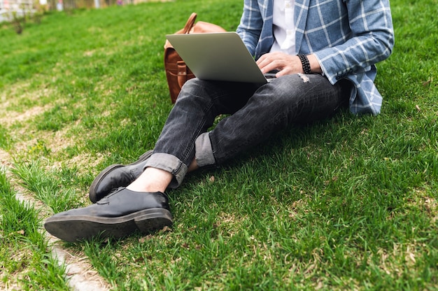 Homem da moda sentado no gramado e trabalhando com um blogueiro freelancer laptop, a vida de uma pessoa moderna