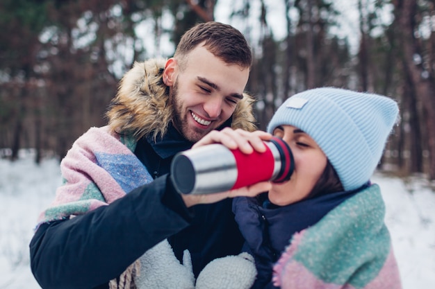 Homem dando a sua namorada chá quente para beber na xícara de garrafa térmica. casal apaixonado andando juntos na floresta de inverno.