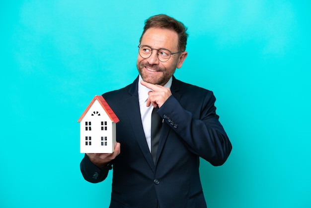 Homem de agente imobiliário de meia idade isolado em fundo azul, olhando para o lado e sorrindo