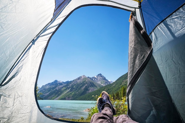 Homem de alpinista de pés relaxando assistindo de uma barraca de acampamento ao ar livre Conceito de estilo de vida de viagem na margem de um lago de montanha montanhas de neve em férias de verão de fundo