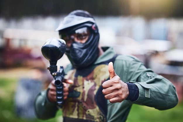 Homem de arma de paintball e retrato com roupas de camuflagem de máscara e polegares para cima para iniciar o jogo de combate ao ar livre Jogos de guerra de cabeça de capacete e aventura para treinamento de tiro ou treinamento de desenvolvimento tático