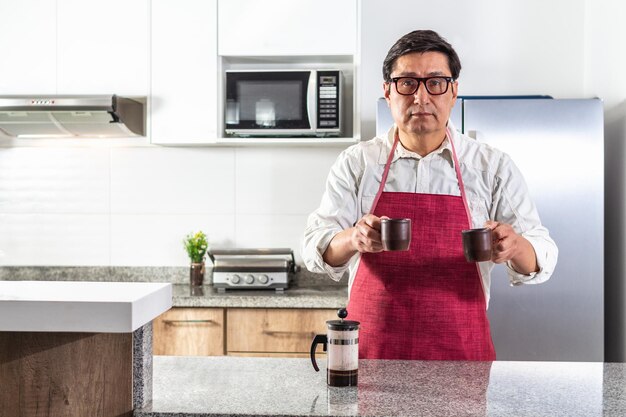 Homem de avental vermelho e óculos entregando duas xícaras de café preparado na prensa francesa no balcão da cozinha