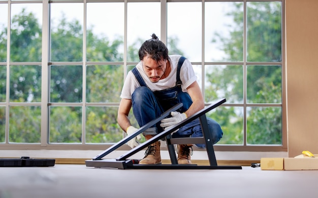 Homem de barba asiático jovem hippie trabalhando como faz-tudo, montando mesa de madeira com equipamentos, conceito para casa diy, hobby e self-service, árvore verde no fundo.