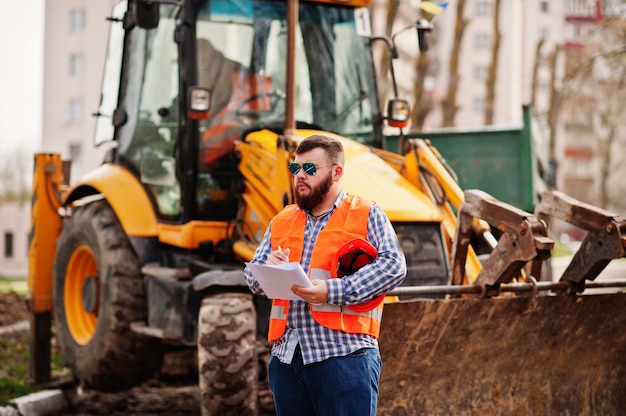 Foto homem de barba brutal trabalhador terno trabalhador da construção civil no capacete de segurança laranja, óculos de sol contra traktor com papel de plano nas mãos.
