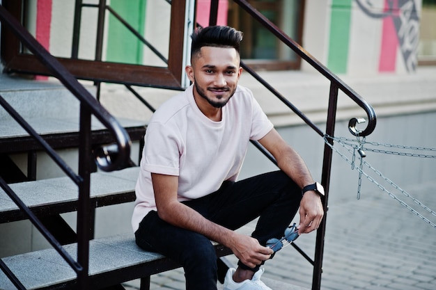 Homem de barba indiana elegante em camiseta rosa modelo Índia sentado em escadas ao ar livre nas ruas da cidade