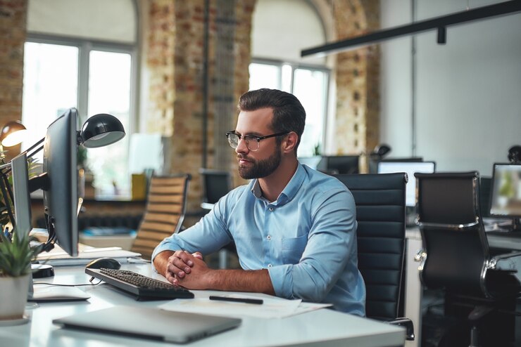Homem de barba jovem e bem-sucedido usando óculos e roupa formal olhando para o monitor do computador