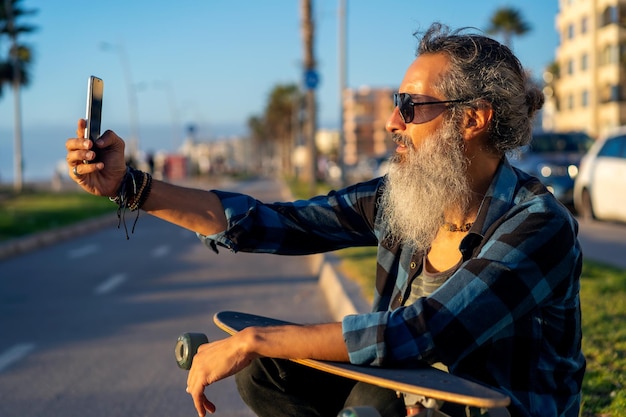 Homem de barba sênior com skate sentado na rua tirando fotos com smartphone ao pôr do solx9