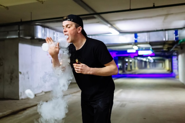 Homem de boné fuma um cigarro eletrônico e libera nuvens de vapor realizando truques de vaping