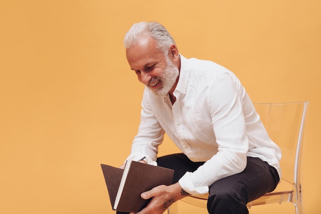 Homem de cabelo branco em poses de camisa com notebook em fundo laranja Cara sorridente barbudo está sentado com livro nas mãos