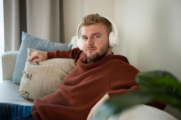 Homem de cabelo cacheado loiro caucasiano jovem ouvindo música com grandes fones de ouvido brancos mãos livres sentado no sofá em casa em fundo branco neutro Estilo de vida de retrato de cara adulto bonitoCopyspace