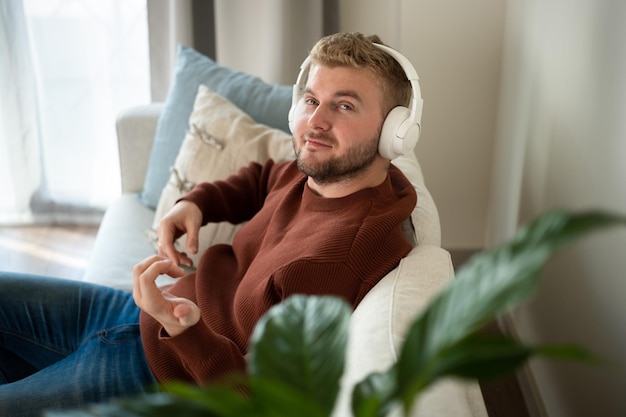 Homem de cabelo cacheado loiro caucasiano jovem ouvindo música com grandes fones de ouvido brancos mãos livres sentado no sofá em casa em fundo branco neutro Estilo de vida de retrato de cara adulto bonitoCopyspace