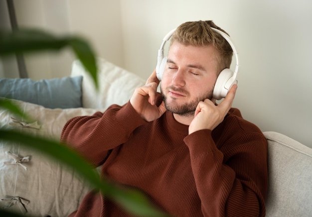 Homem de cabelo cacheado loiro caucasiano jovem ouvindo música com grandes fones de ouvido brancos mãos livres sentado no sofá em casa em fundo branco neutro Estilo de vida de retrato de cara adulto bonitoCopyspace