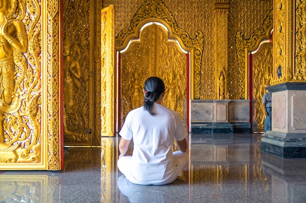 Homem de cabelo comprido asiático relaxa a meditação com todo o traje branco sentado em frente ao papel de parede dourado do budista no templo, Tailândia.