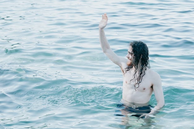 Foto homem de cabelo comprido dentro de água cristalina saudando alguém na praia, água cristalina, conceito de férias