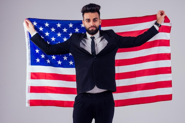 Homem de cabelo preto bonito jovem árabe em terno de negócios elegante segurando a bandeira americana no estúdio isolar o fundo brancoconceito do Dia da Independência