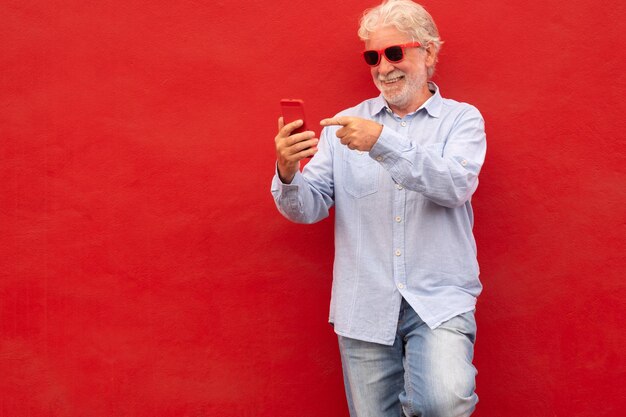 Homem de cabelos brancos bonito sênior vestindo camisa casual e óculos sobre fundo vermelho segurando o telefone no bate-papo por vídeo pessoa feliz gesticulando com a mão