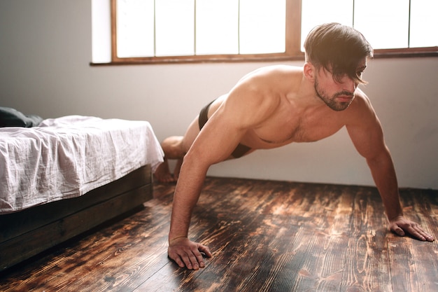 Homem de cabelos escuros barbudo nu fazendo exercícios de flexão de manhã em casa.