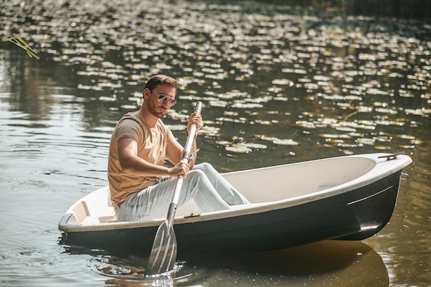 Homem de cabelos escuros focado sério em óculos de sol sentado sozinho no barco remando com um remo