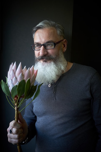 Homem de cabelos grisalhos com óculos e barba tem um protea rosa nas mãos