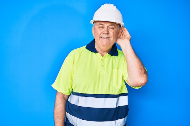 Homem de cabelos grisalhos sênior vestindo camiseta reflexiva de trabalhador e capacete sorrindo com a mão sobre a orelha ouvindo e ouvindo boatos ou fofocas sobre o conceito de surdez