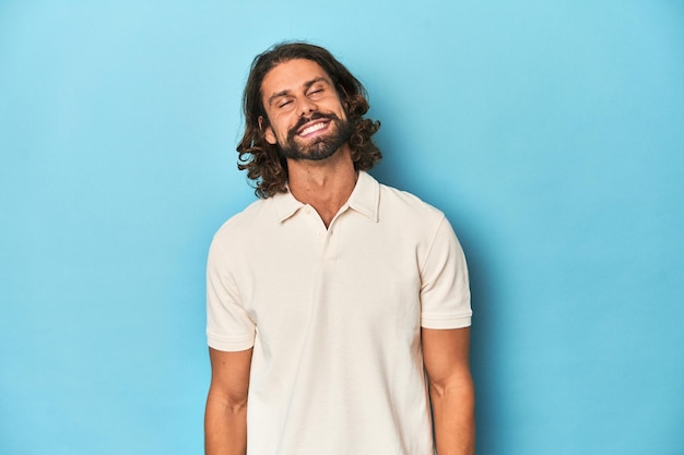 Foto homem de cabelos longos com um estúdio azul branco ri-se e fecha os olhos sente-se relaxado e feliz