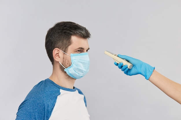 Homem de camisa azul e máscara de proteção no rosto olhando para um termômetro eletrônico que o médico segura com luvas azuis e mede sua temperatura