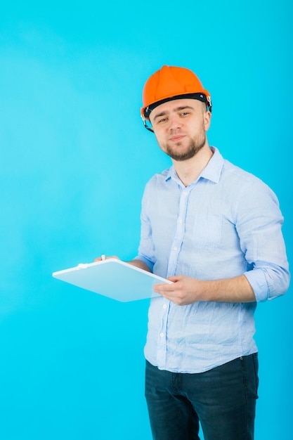 homem de camisa azul em um capacete protetor laranja e um notebook branco fica em um fundo azul