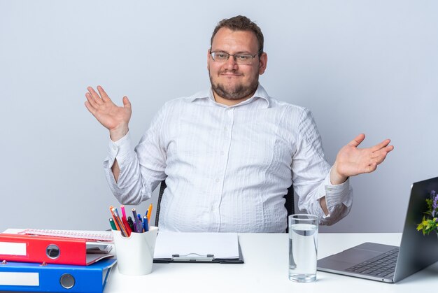 Homem de camisa branca de óculos olhando para a frente feliz e satisfeito abrindo os braços para os lados, sentado à mesa com pastas de escritório do laptop e prancheta sobre a parede branca trabalhando no escritório
