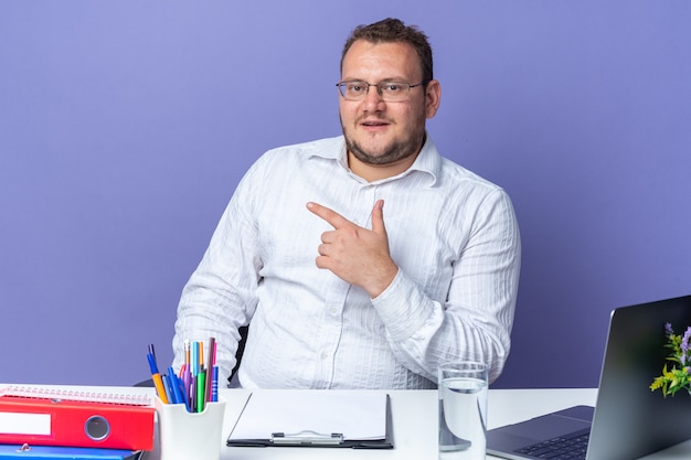 Homem de camisa branca de óculos sorrindo confiante apontando com o dedo indicador para o lado sentado à mesa com laptop e pastas de escritório sobre a parede azul trabalhando no escritório