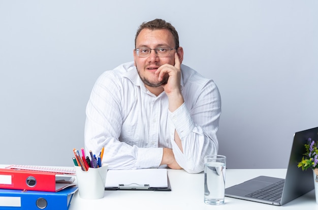 Homem de camisa branca, usando óculos, olhando para cima, pensando positivo, sorrindo, sentado à mesa com pastas de escritório do laptop e prancheta sobre a parede branca, trabalhando no escritório