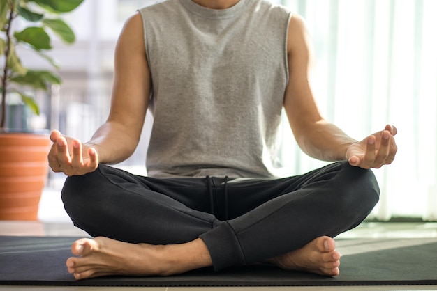 Homem de camisa cinza sem mangas e calça preta sentado no colchonete praticando meditação sentado em uma pose de lótus sozinho dentro de casa durante a quarentena devido à disseminação do coronavírus