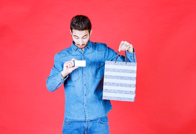 Homem de camisa jeans, segurando uma caixa de compras listrada e apresentando seu cartão de visita.