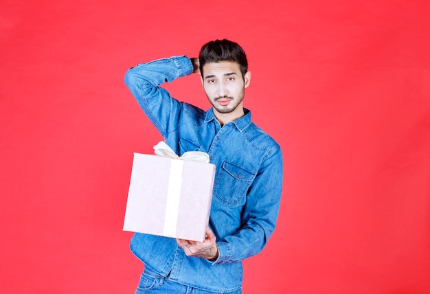 Homem de camisa jeans segurando uma caixa de presente roxa amarrada com fita branca e parece confuso e pensativo.