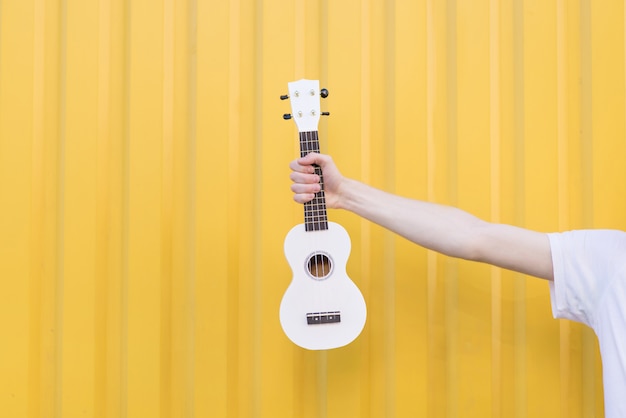 Homem de camiseta branca segura um ukulele contra uma parede amarela