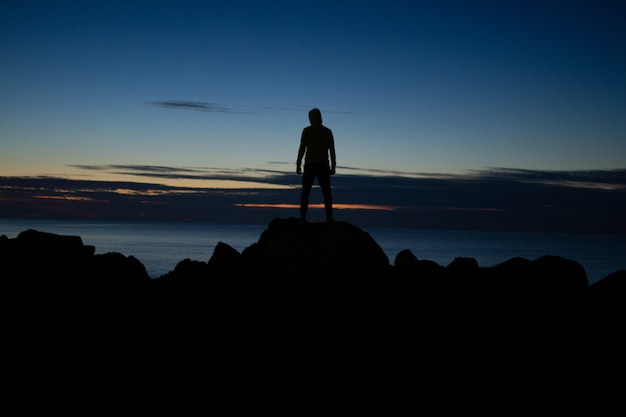 Homem de capuz em pé sobre as rochas no fundo do mar no entardecer