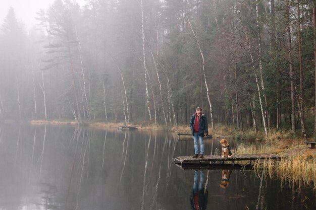 Homem de casaco azul ficar com o cachorro no cais na margem do lago. Macho andando com o retriever de pedágio marrom do pato da Nova Escócia. Nevoeiro acima da água e floresta no fundo. Foco seletivo, copie o espaço.