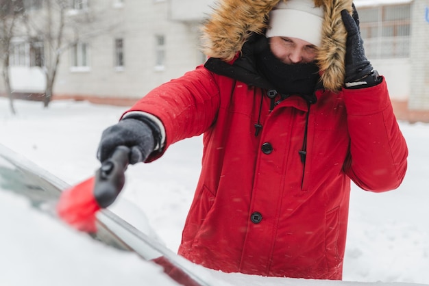 Homem de casaco de inverno vermelho com capô de pele limpando carro após tempestade de neve
