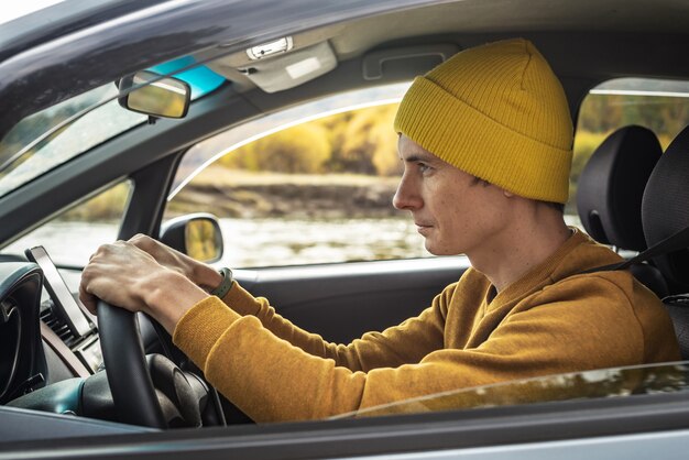 Homem de chapéu amarelo e camisola está dirigindo um carro ao longo do rio e a bela floresta de outono. Conceito de natureza, viagens e clima de outono