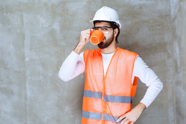 Homem de engenheiro com capacete branco bebendo de uma caneca laranja.