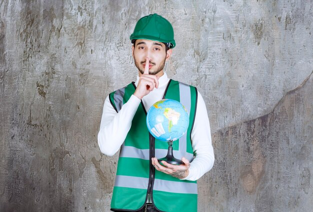 Homem de engenheiro de uniforme amarelo e capacete segurando um globo do mundo e pedindo silêncio.