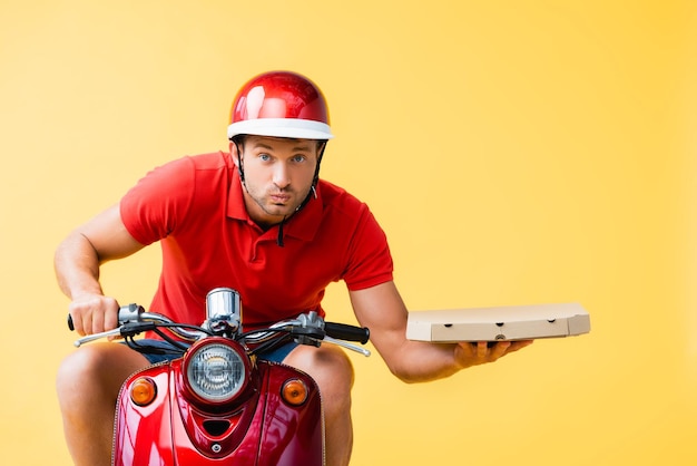 Homem de entrega concentrado no capacete andando de scooter vermelho e segurando a caixa de pizza isolada em amarelo