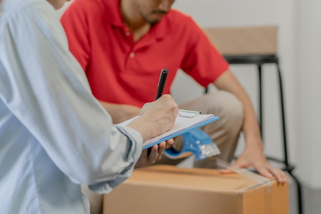 Foto homem de entrega da empresa de carga, embalagem masculina por máquina de fita, caixa de papelão de vedação. proprietária mulher asiática de negócios verificando coisas da lista na área de transferência no armazém. movendo-se, preparando-se em nova casa, realocação.