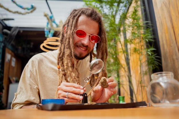 Homem de estilo hippie examina sob uma lupa as articulações e botões de maconha medicinal