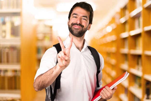 Homem de estudante, fazendo o gesto de vitória na biblioteca desfocada