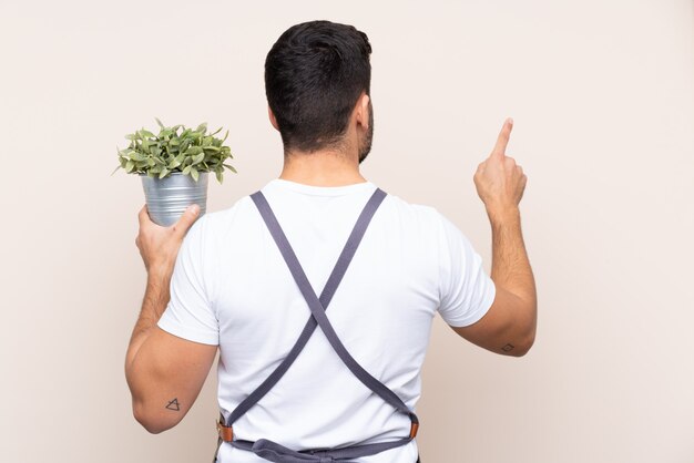 Homem de jardineiro segurando uma planta sobre parede isolada, apontando para trás com o dedo indicador