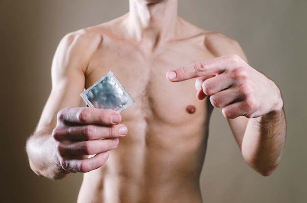 homem de jeans preto está segurando uma camisinha, enquanto a outra mão está apontando para um contraceptivo com o dedo indicador.