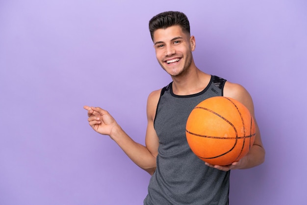 Homem de jogador de basquete jovem caucasiano isolado no fundo roxo, apontando o dedo para o lado