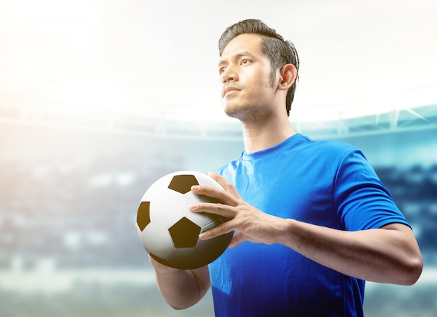 Homem de jogador de futebol asiático em jersey azul segurando a bola no campo de futebol