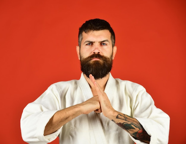 Homem de karatê com rosto confiante de uniforme. homem com barba no quimono branco sobre fundo vermelho. conceito de esportes orientais. mestre de taekwondo coloca o punho na palma da mão em saudação tradicional