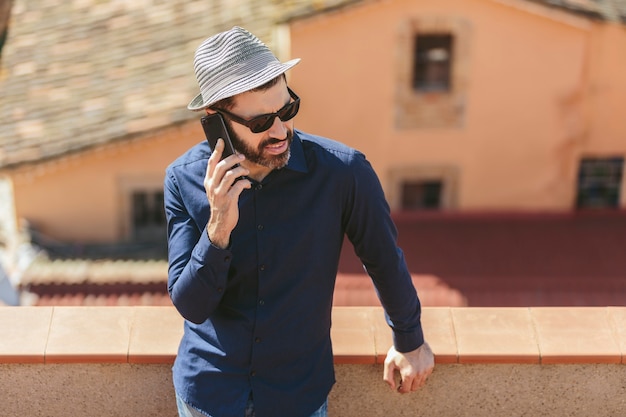 Homem de meia-idade atraente com chapéu e óculos escuros, falando com um telefone inteligente em pé na varanda.